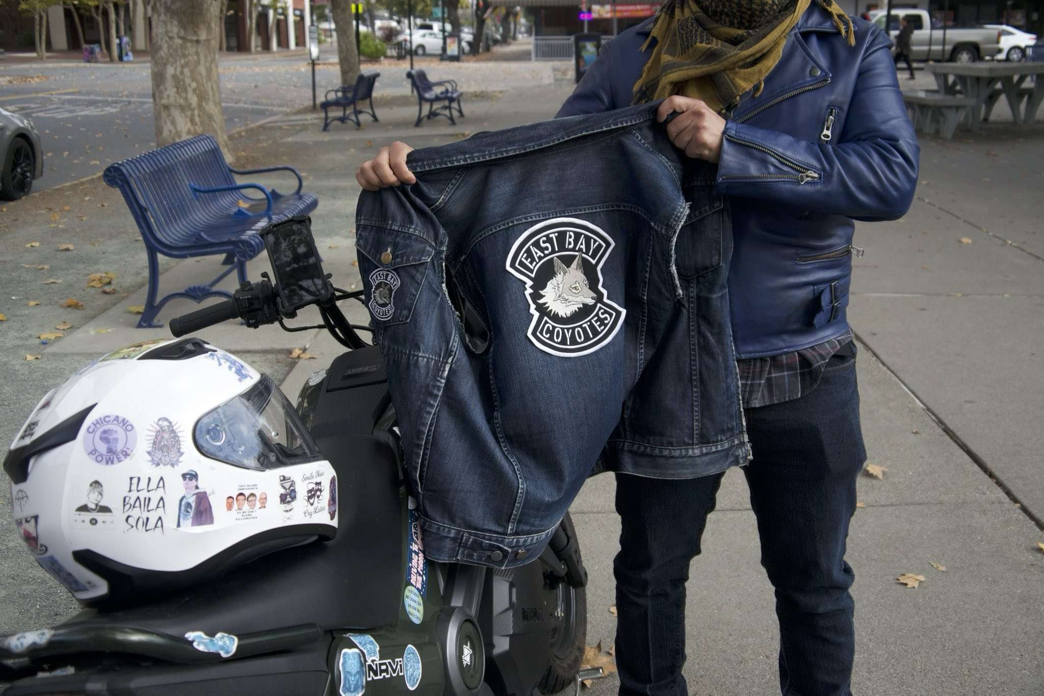 A person in jeans an a blue leather jacket holds a denim vest with a motorcycle club patch on the back that says "East Bay Coyotes" with a coyote in the middle