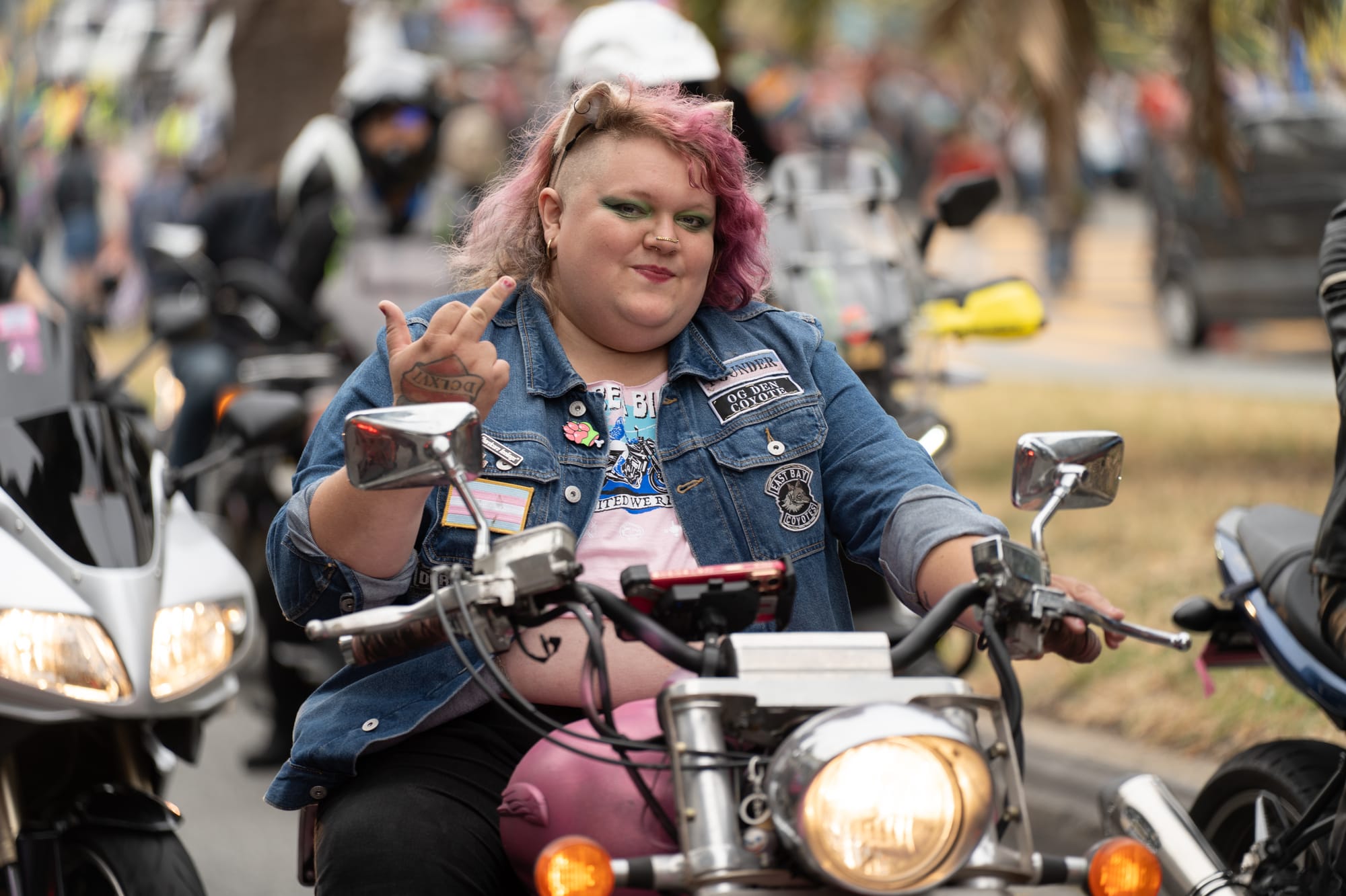 A person with pink hair and cat ears flips off the camera while riding a pink motorcycle