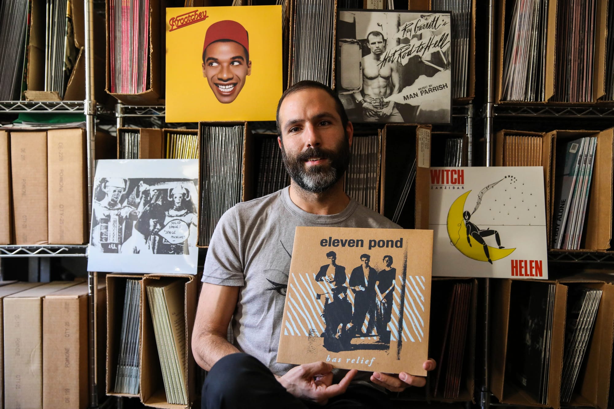 a white man with dark hair and a beard sits in front of vinyl holding a record that reads 'eleven pond, bas relief'