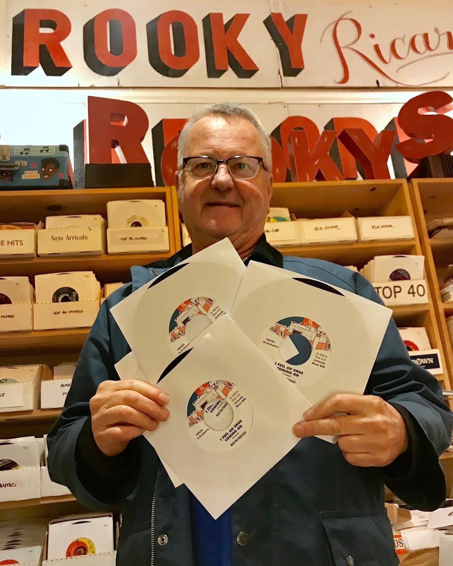 an older white man with glasses holds 45s that read 'i feel an urge coming on' by nick waterhouse, with lots of records behind him