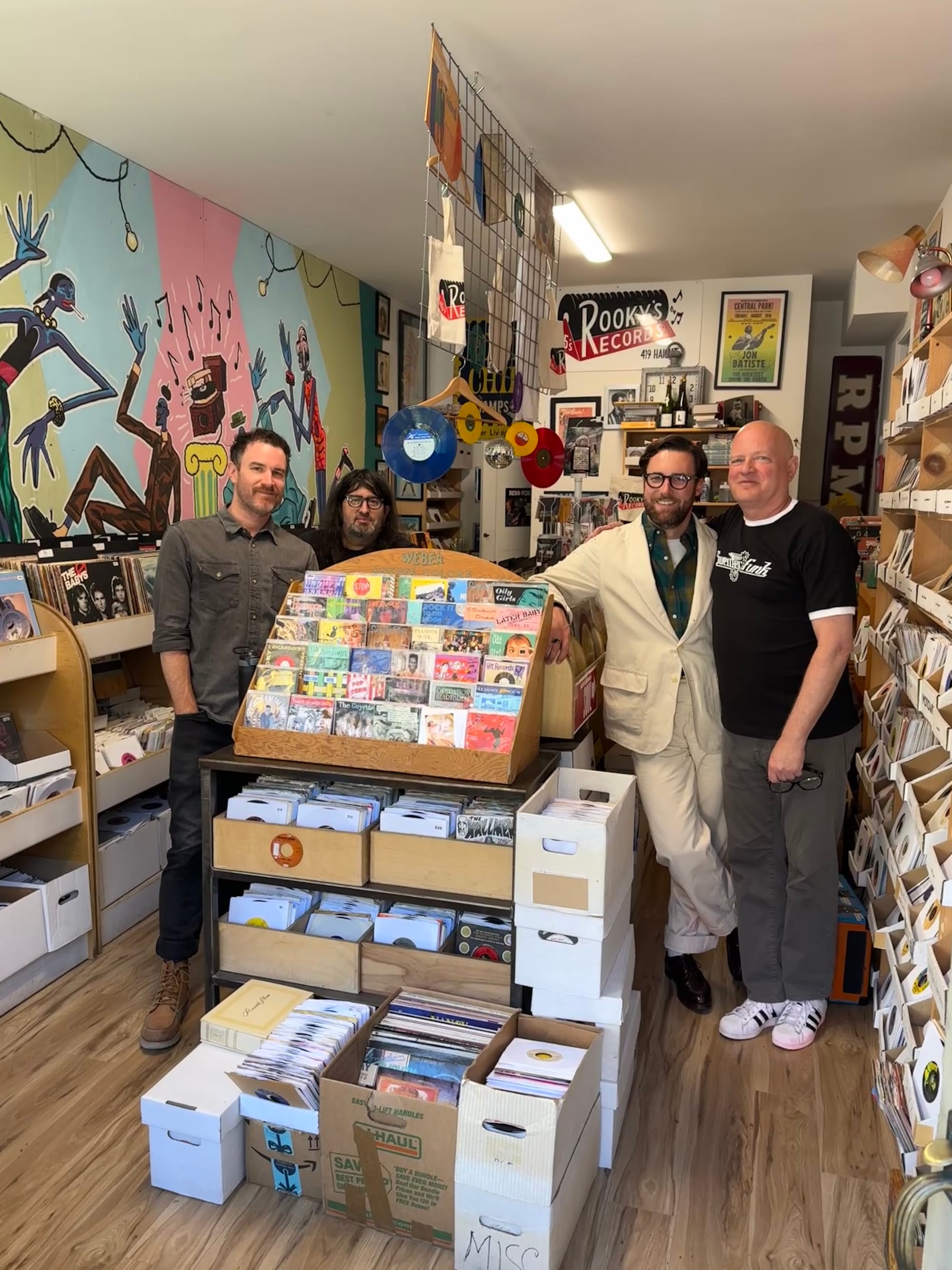 four men in their 30s to 50s stand in a record shop around piles of boxes of vinyl