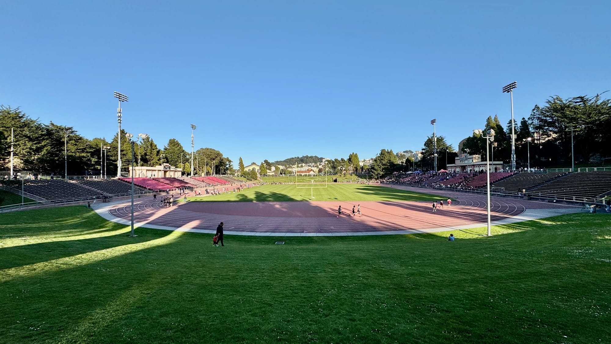 A person walking around the running track surrounding a football field. The grass in the foreground is covered in shadow. 
