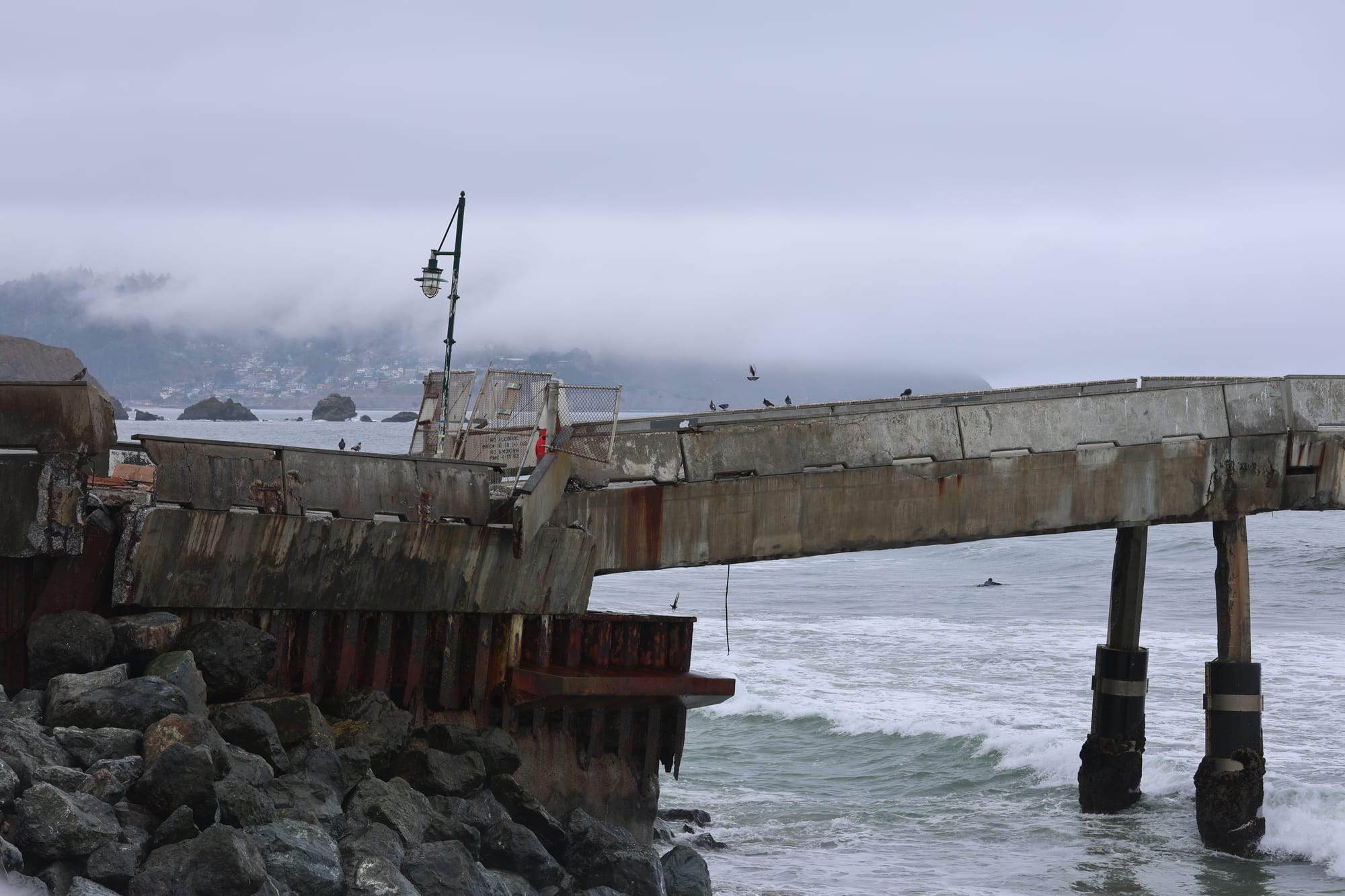 A long concrete pier is cracked at its base on the rocky shore