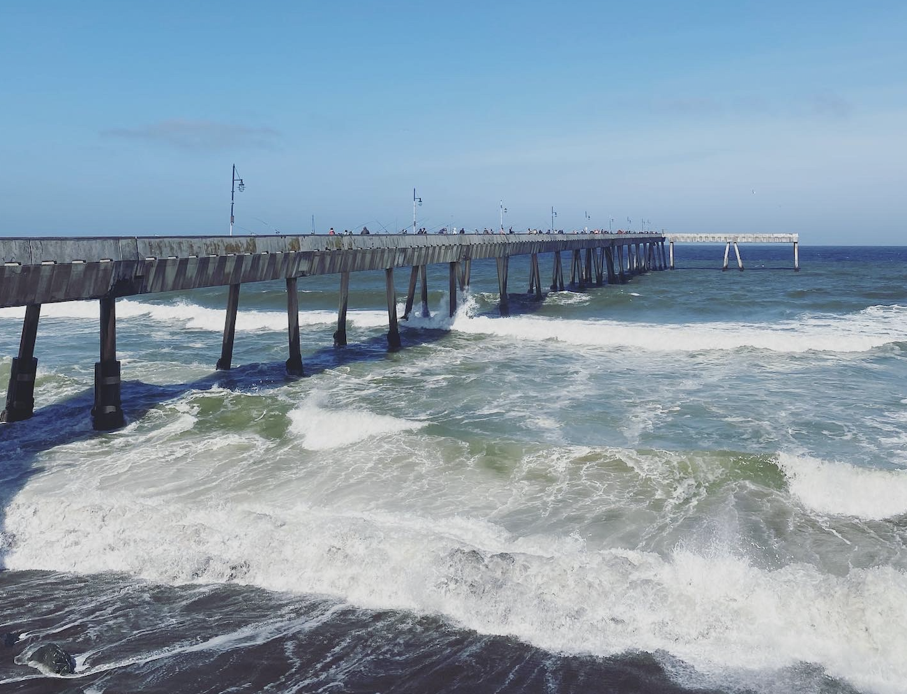 A photo of Pacifica’s concrete pier extending way into the ocean while waves crash beneath it.