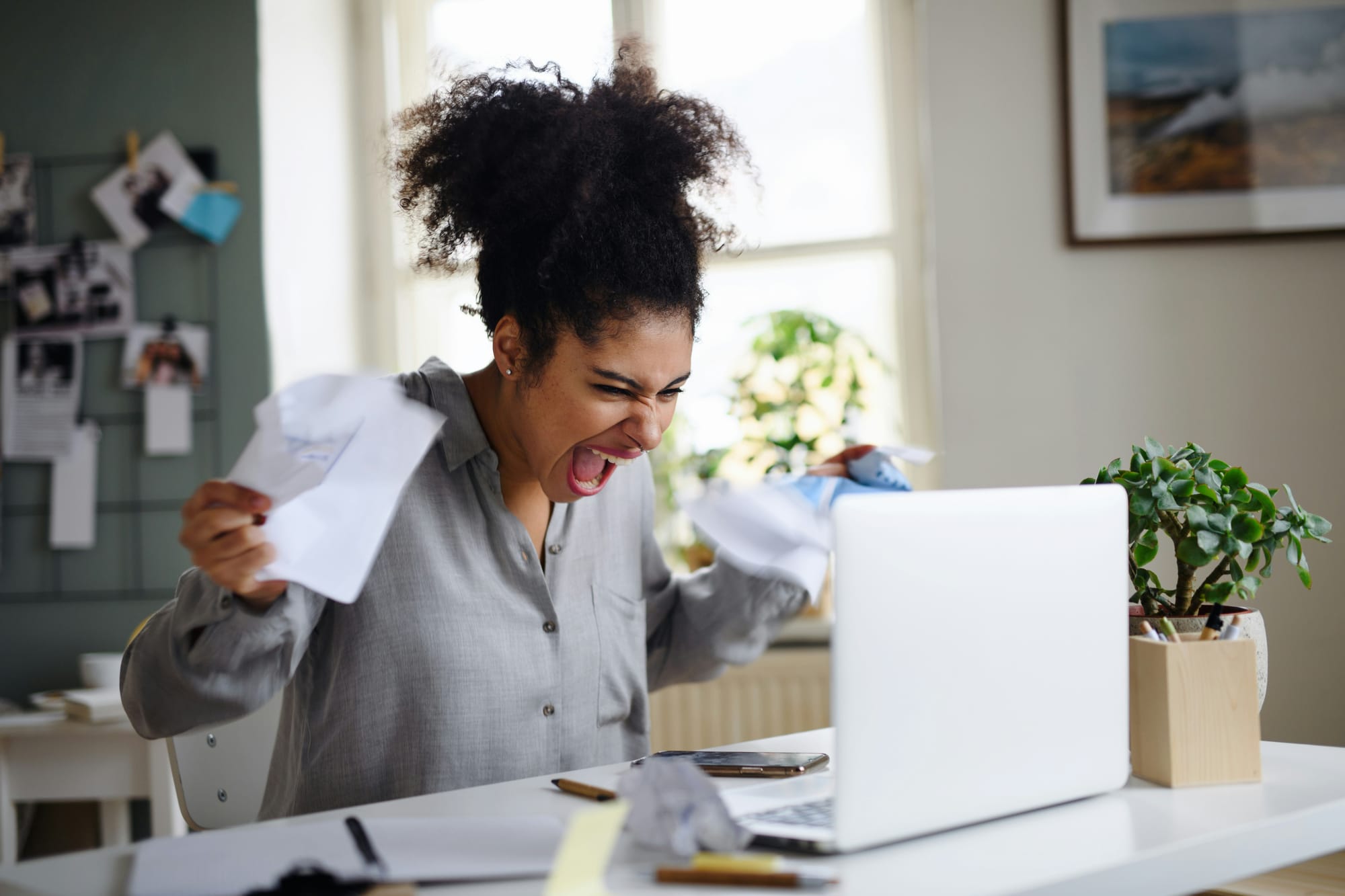 a woman in a grey button up shirt yells at her laptop while holding paper