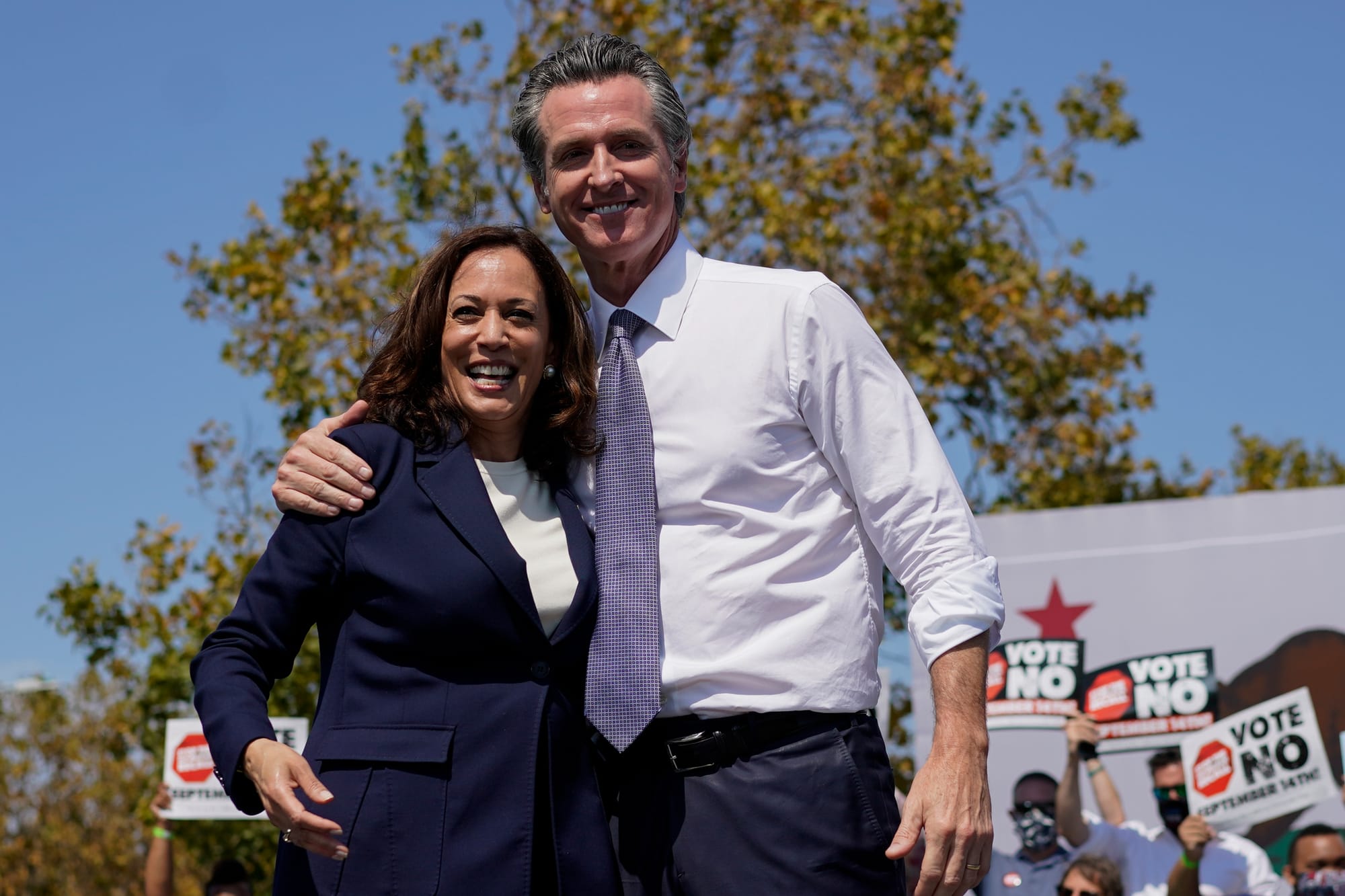 Kamala Harris and Gavin Newsom, two California politicians, a Black woman and a white man, dressed formally, grinning outside at a rally