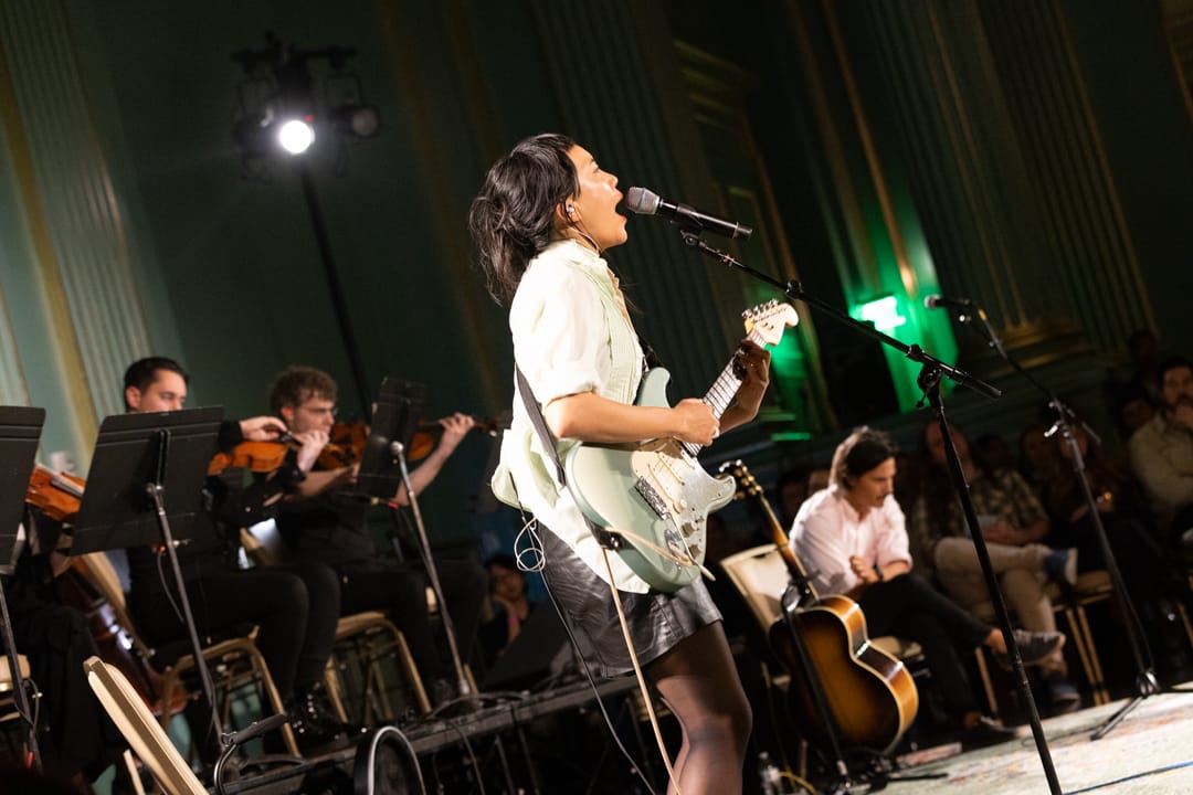 a young woman with black hair in a white shirt and black skirt plays guitar and sings into a microphone with violin players behind her in a green beaux arts room in SF
