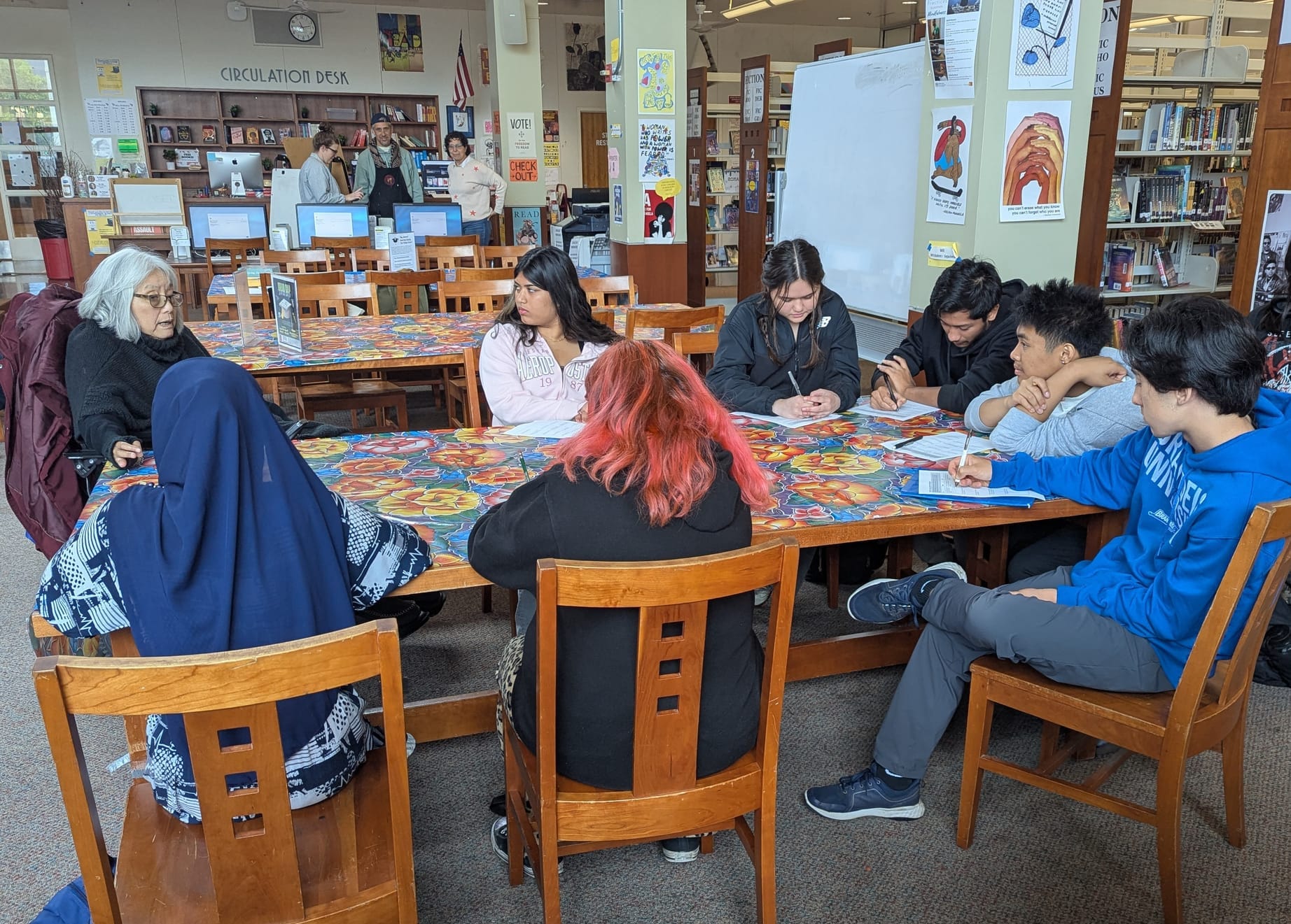 A woman with a grey bob sitting at a table in a library speaking to a group of teenagers.