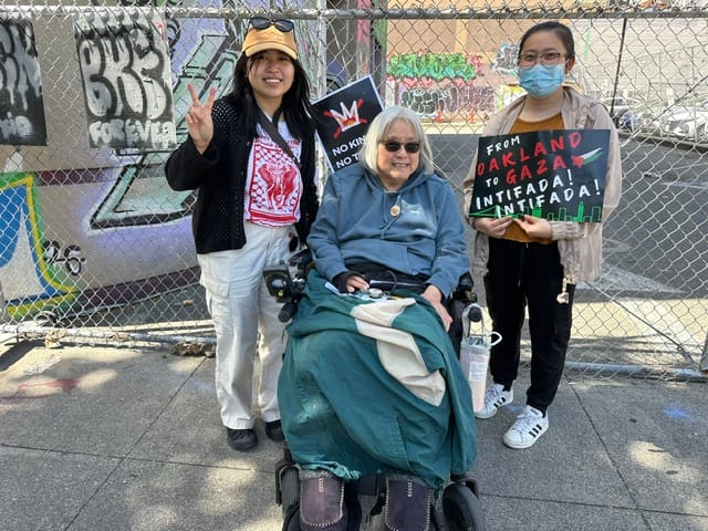 A woman sitting in a wheelchair smiling in front of two people holding "No Kings" and "From Oakland to Gaza Intifada Intifada" signs