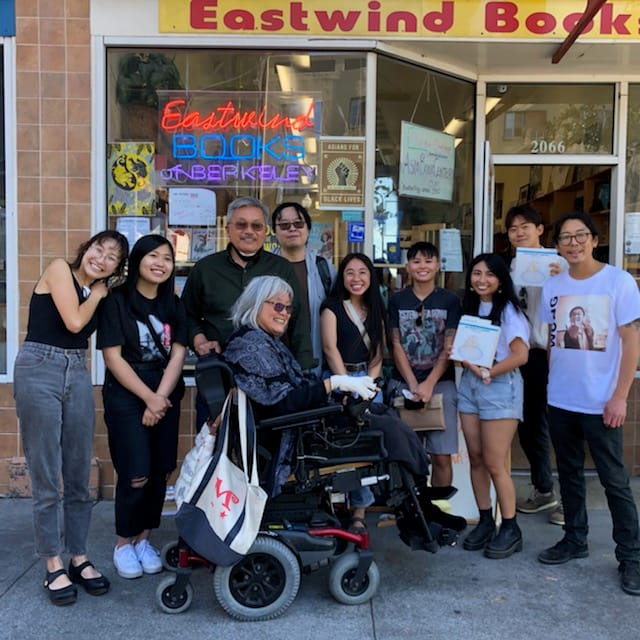 A woman in a wheelchair smiling in front of a group of people standing outside of Eastwind Books