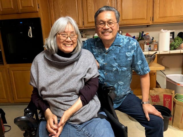 A woman with a grey bob (sitting in a wheelchair) and a man with a mustache (crouching) smiling in a home kitchen