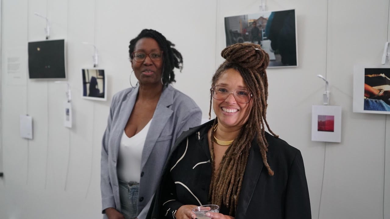 two women stand in front of a wall with art on it at a gallery opening, smiling, dressed nicely