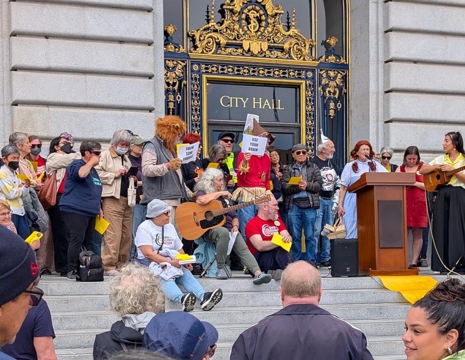 a protest at san francisco city hall with about 20 people on the steps, someone holding a guitar, and some people dressed as characters from The Wizard of Oz, as supporters watch