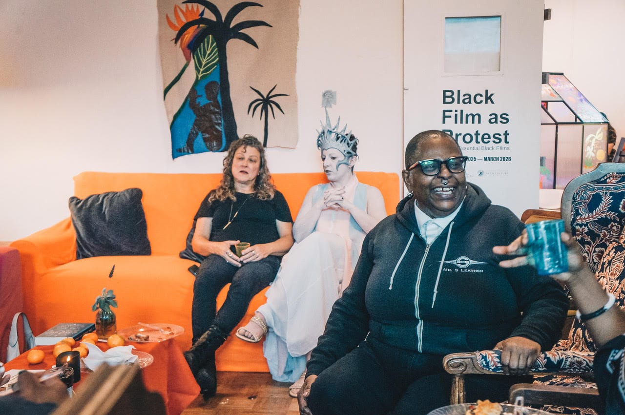 three people, one in white makeup with an elaborate headdress on, sit in a gallery room talking and laughing. Two of them sit on an orange couch, another person sits in a chair in the foreground, laughing at something a person out of frame is saying