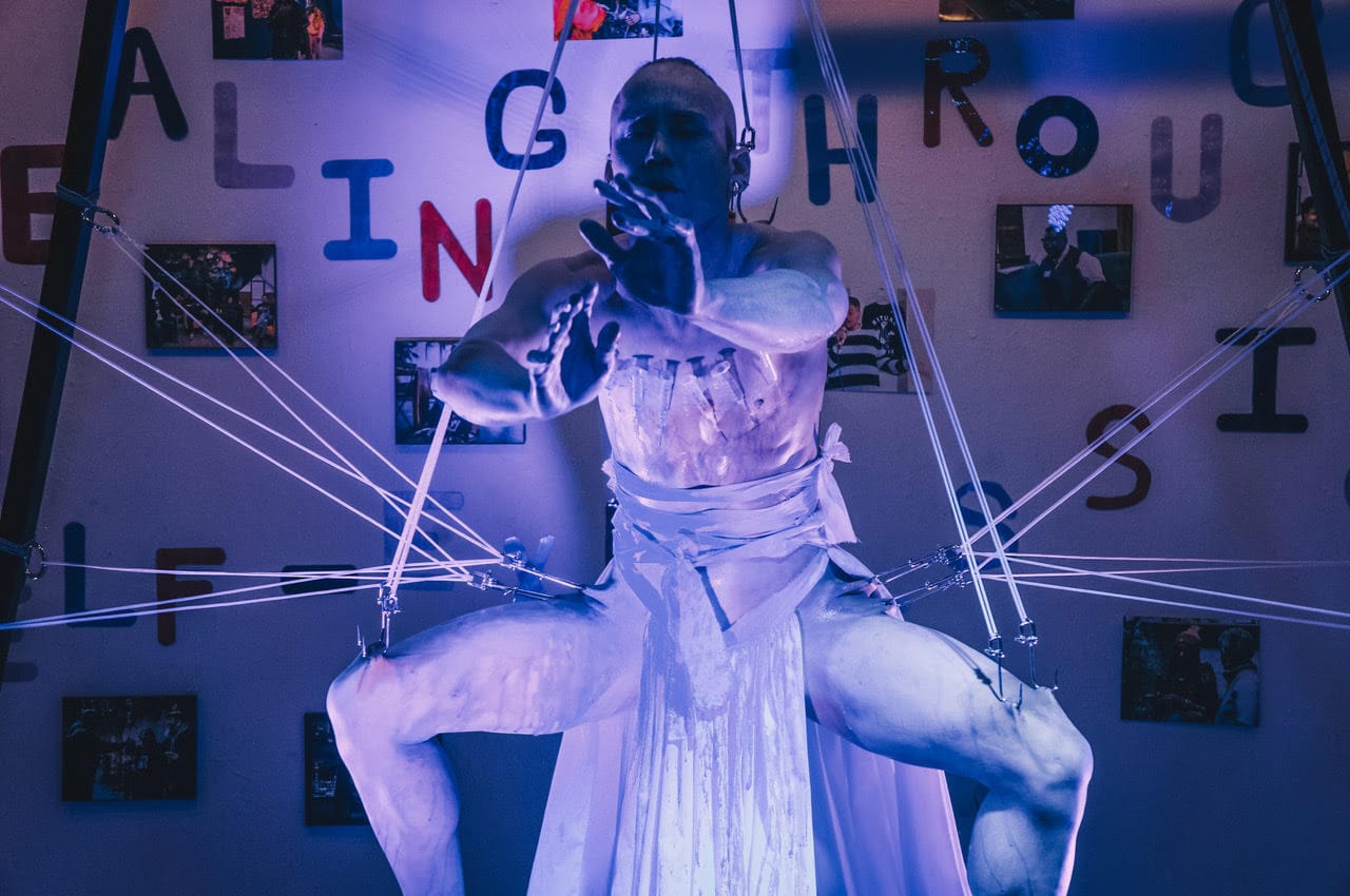 a suspension artist in white makeup performs in a gallery lit dimly in purple with letters and photos on the back wall