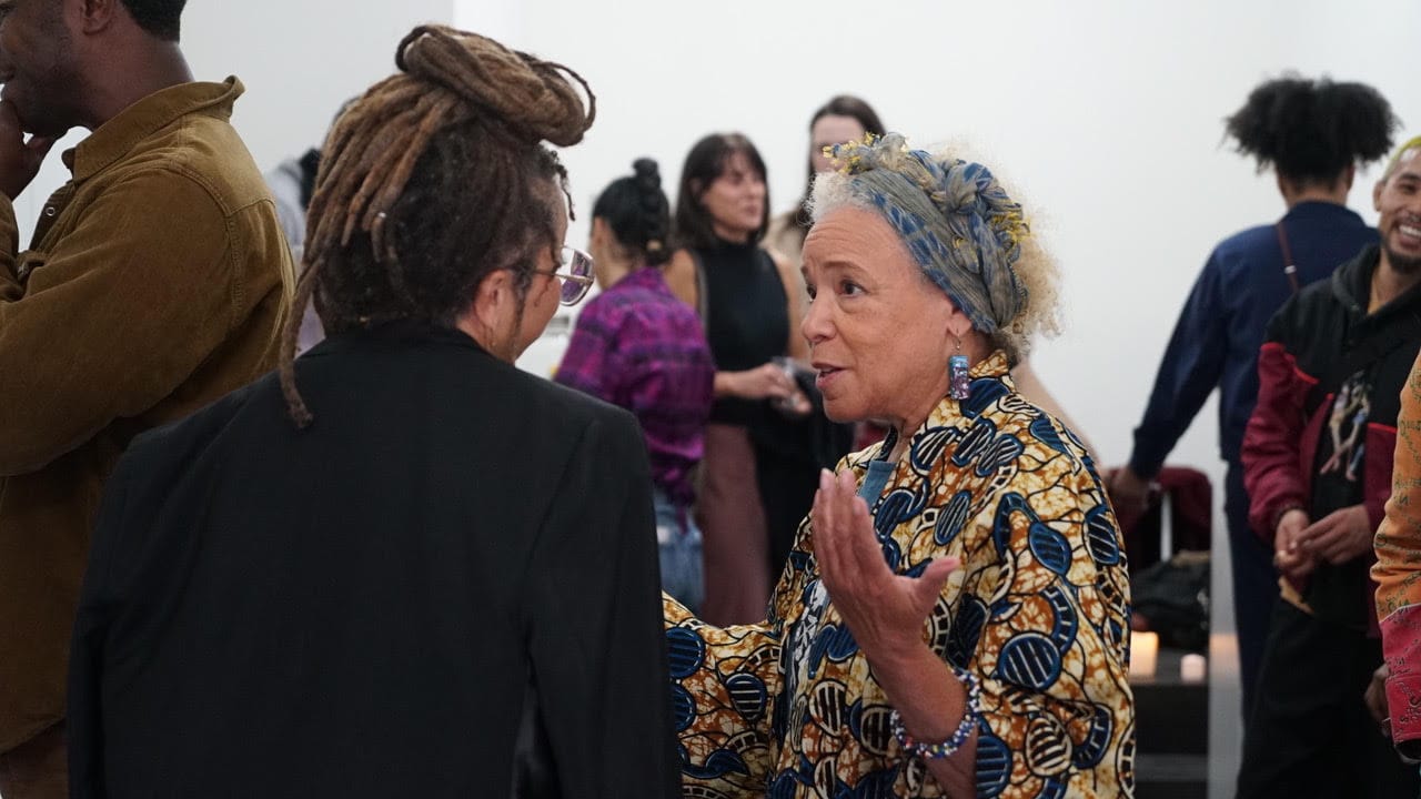 two women, one with her back to the camera, another in a colorful shirt, talk to each other surrounded by others at a gallery opening