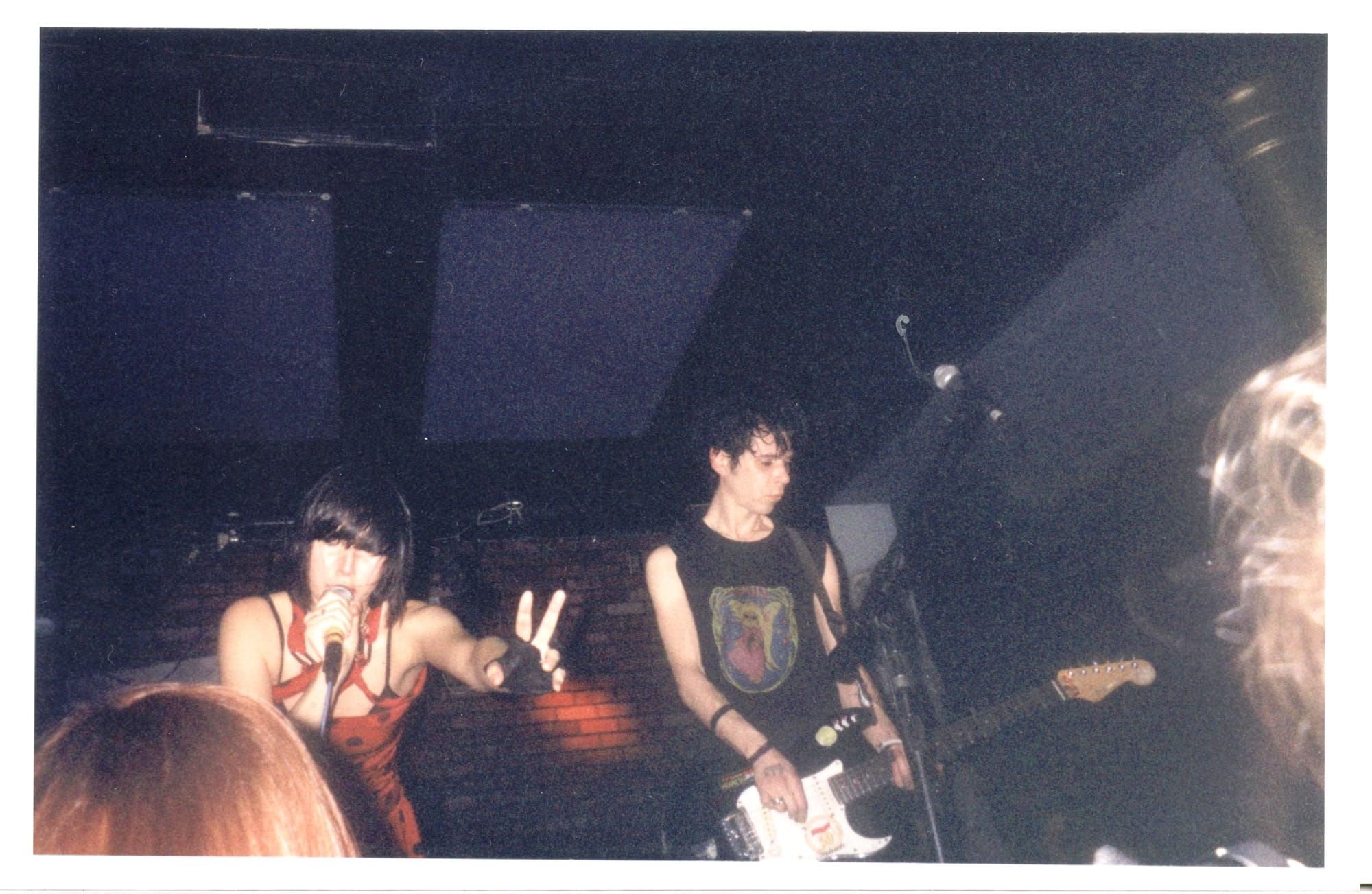 a polaroid of a woman with black hair and another person with black hair playing on a stage in front of a brick wall