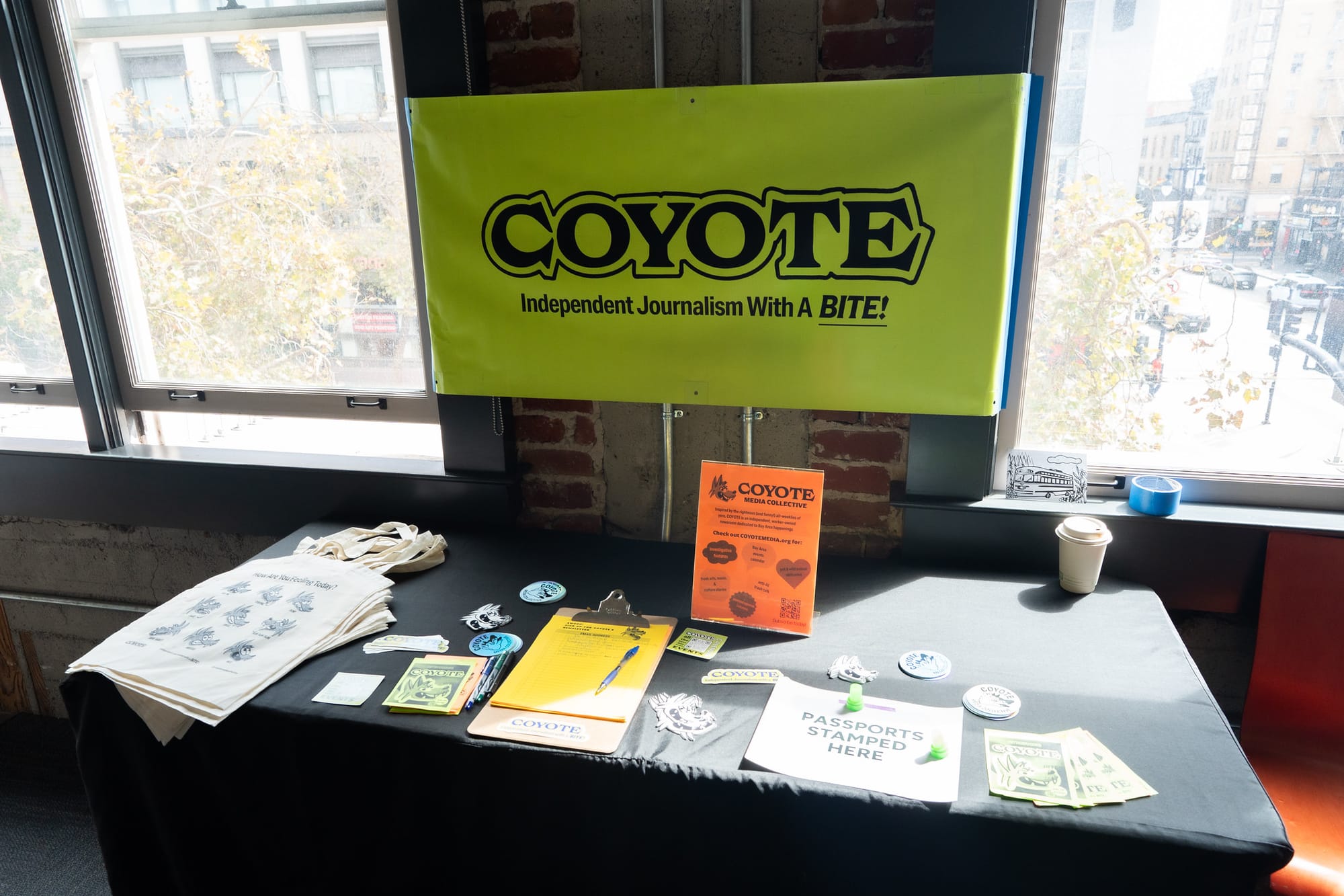 Neon green sign that reads "COYOTE" hanging on the wall behind a table display of flyers, stickers and tote bags.
