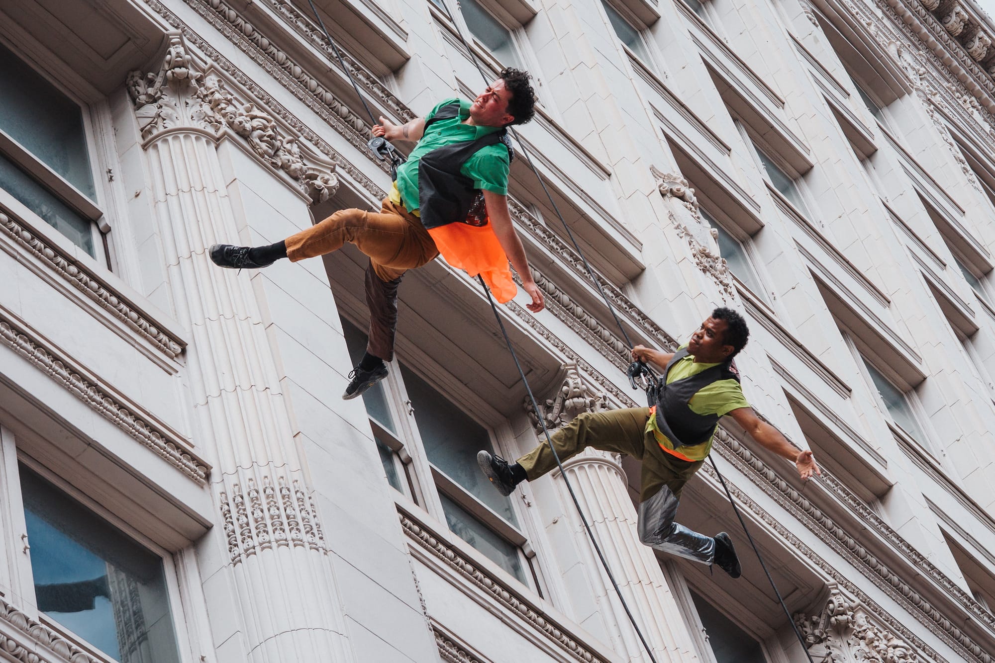 Two people tied to cables dance perpendicular to the side of a large building with their arms outstretched.