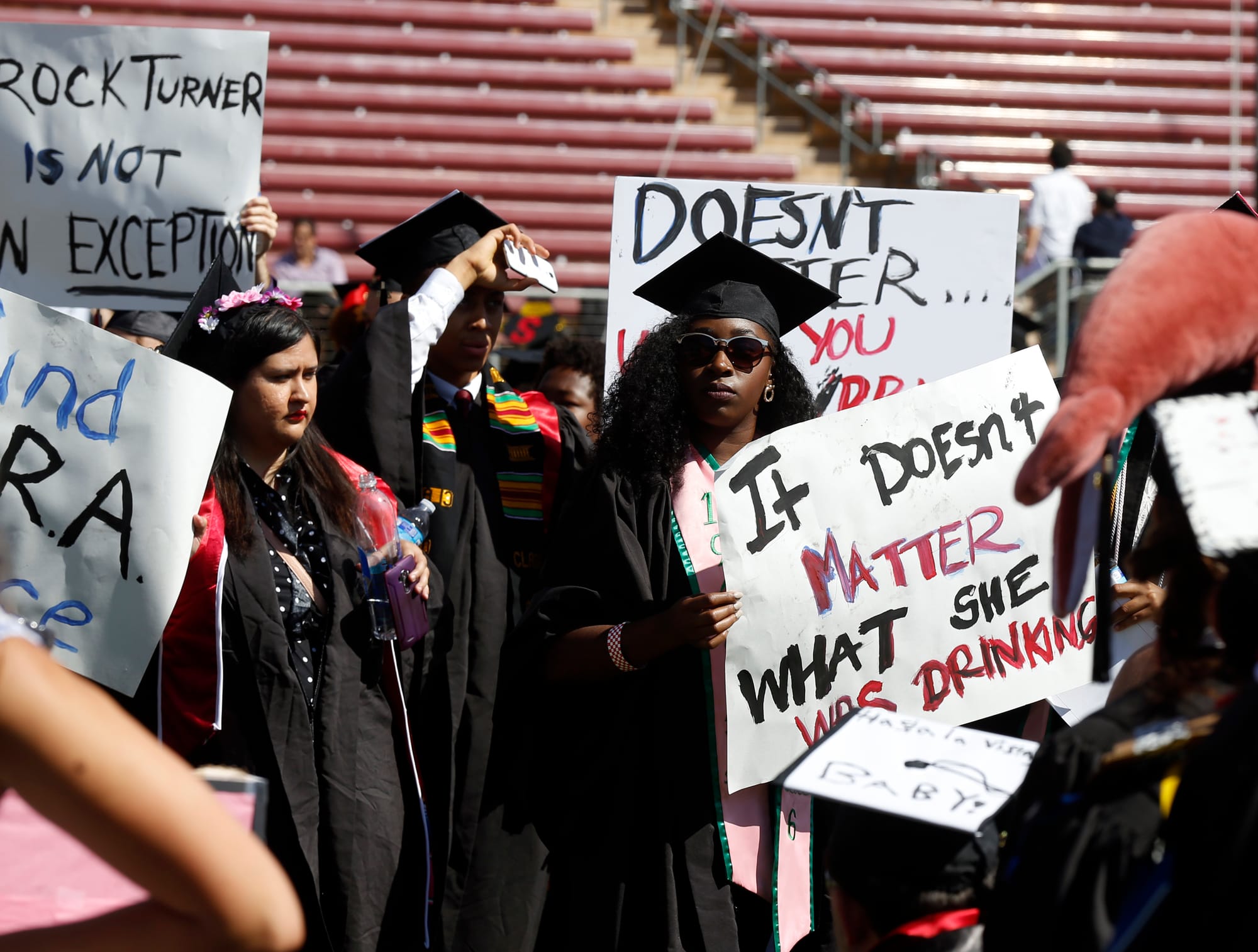 people in graduation gowns hold signs that read 'it doesn't matter what she was drinking' at a protest at a stanford graduation