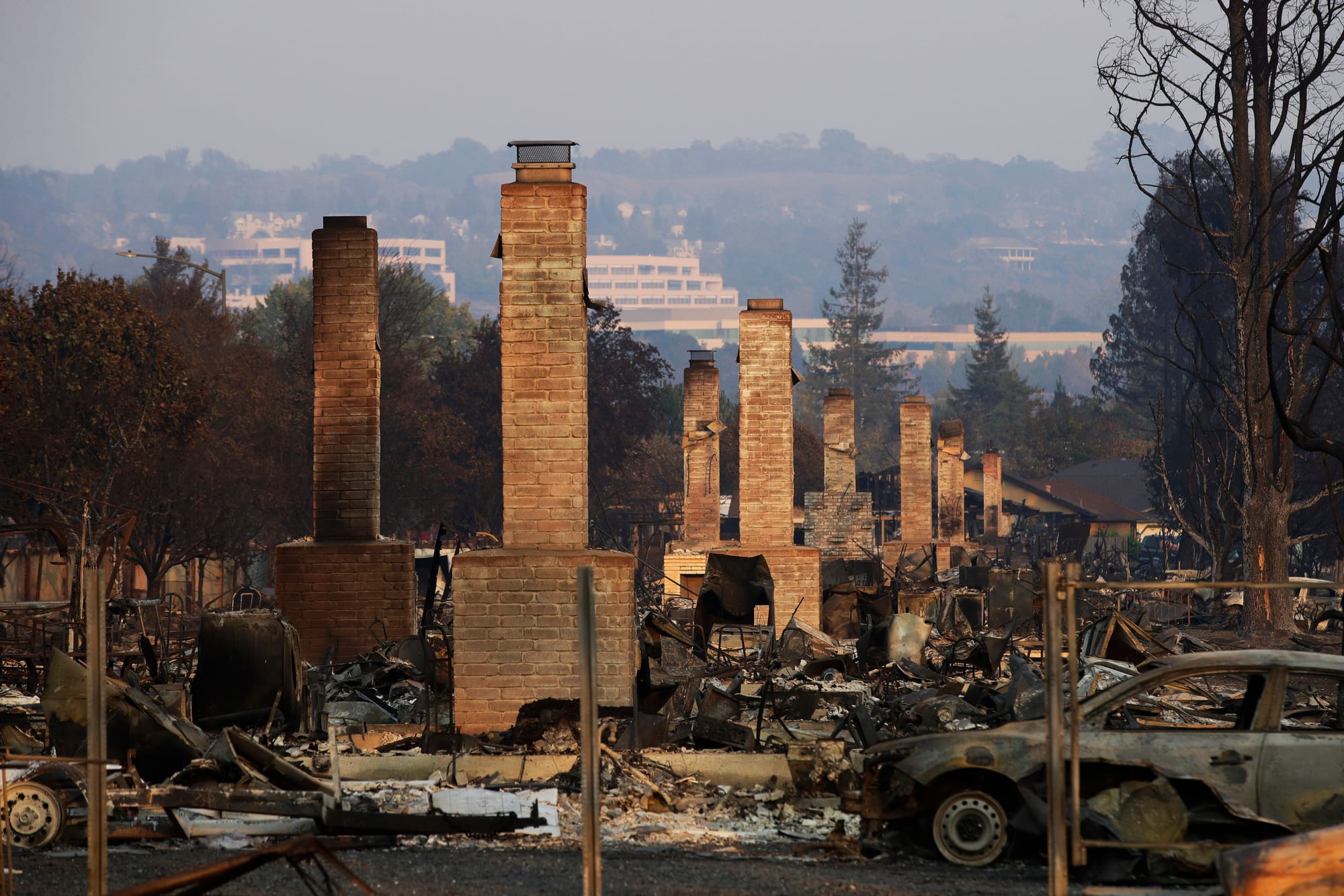 Chimneys are all that remain of an area devastated by fire, with a burnt-out car in the foreground