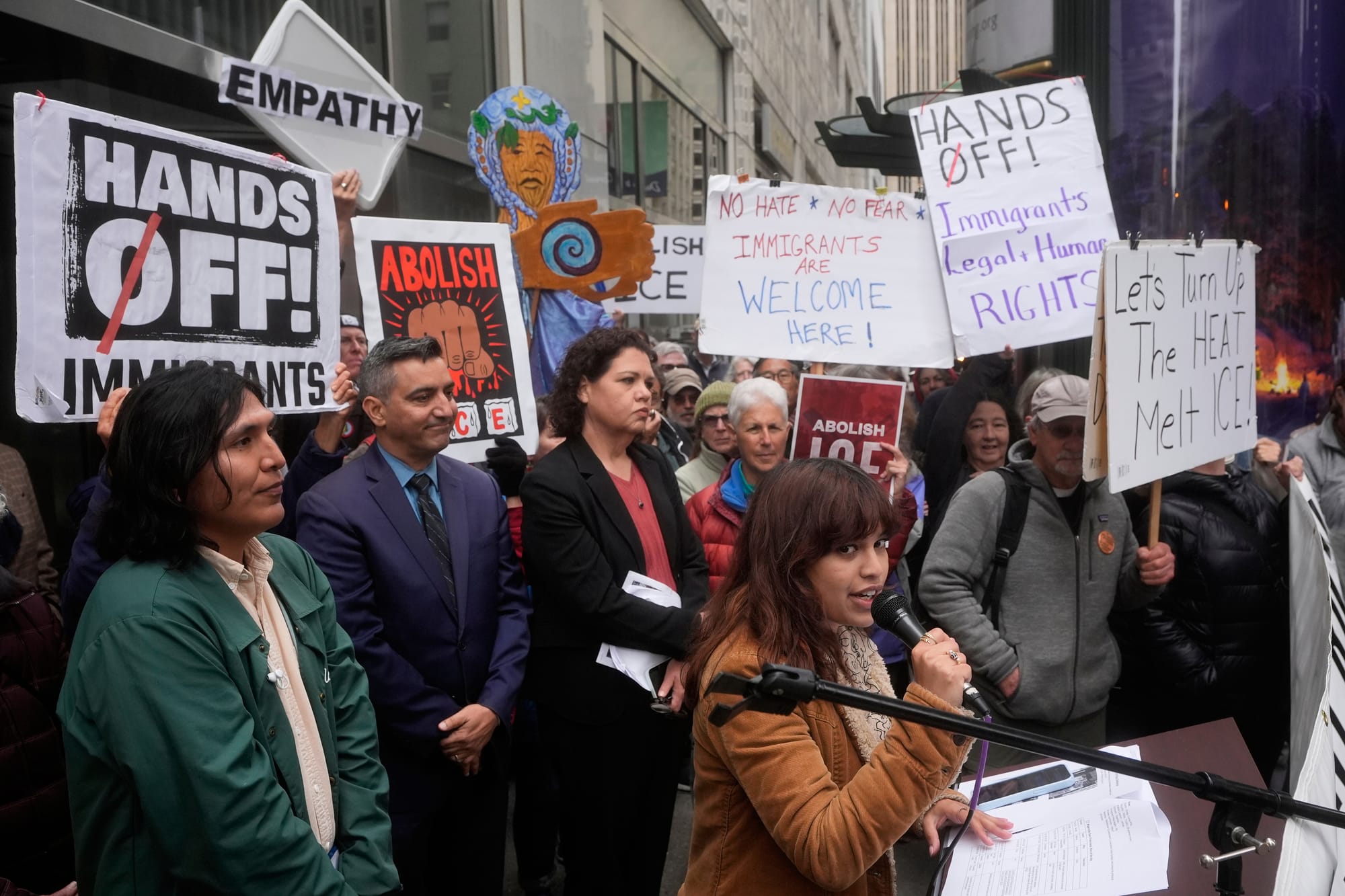 Group of people standing behind a person speaking while holding signs like "Hands Off Immigrants" and "No Hate No Fear Immigrants Are Welcome Here"