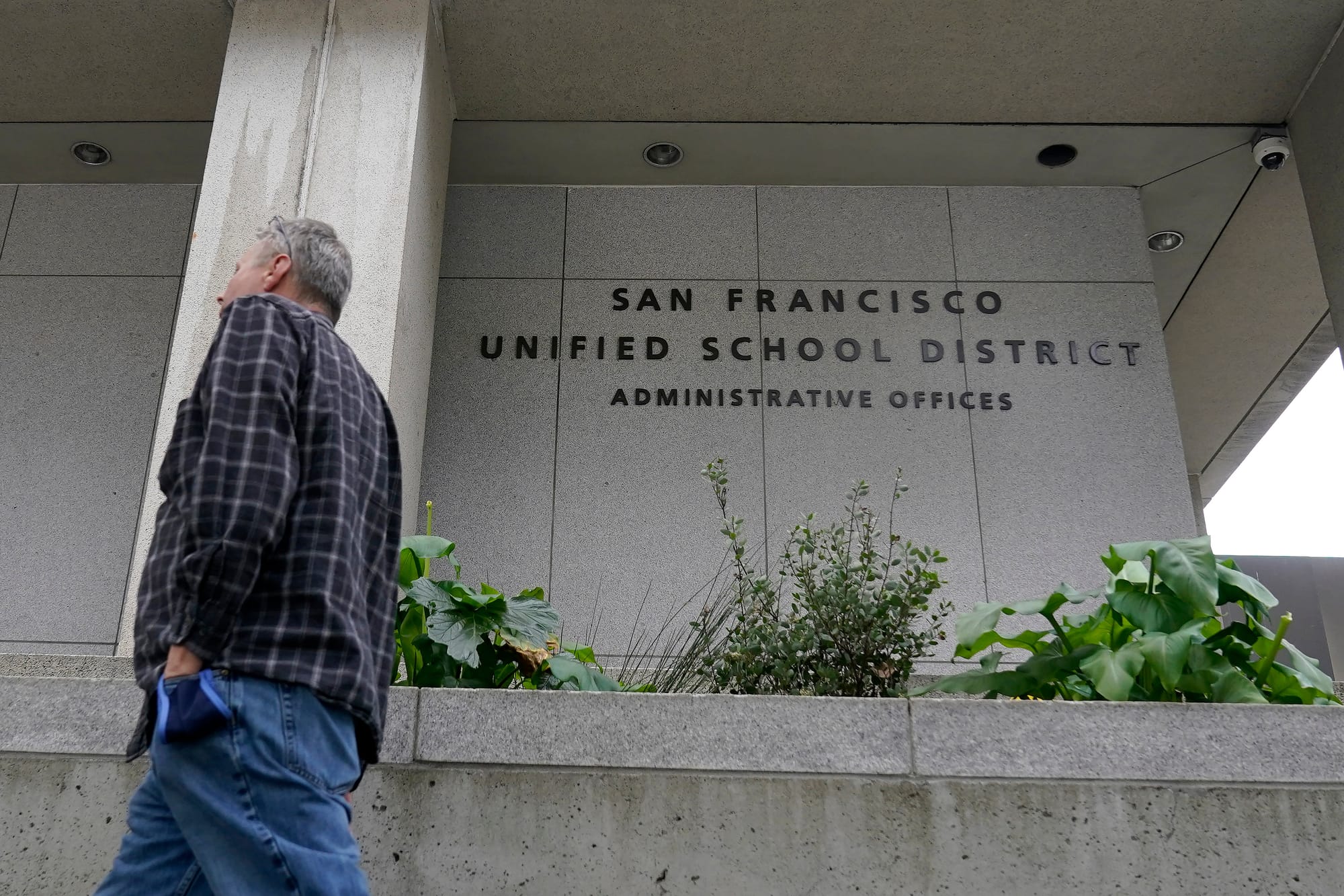 a person in jeans and a flannel shirt walks by a concrete building with letters that read 'san francisco unified school district administrative offices'