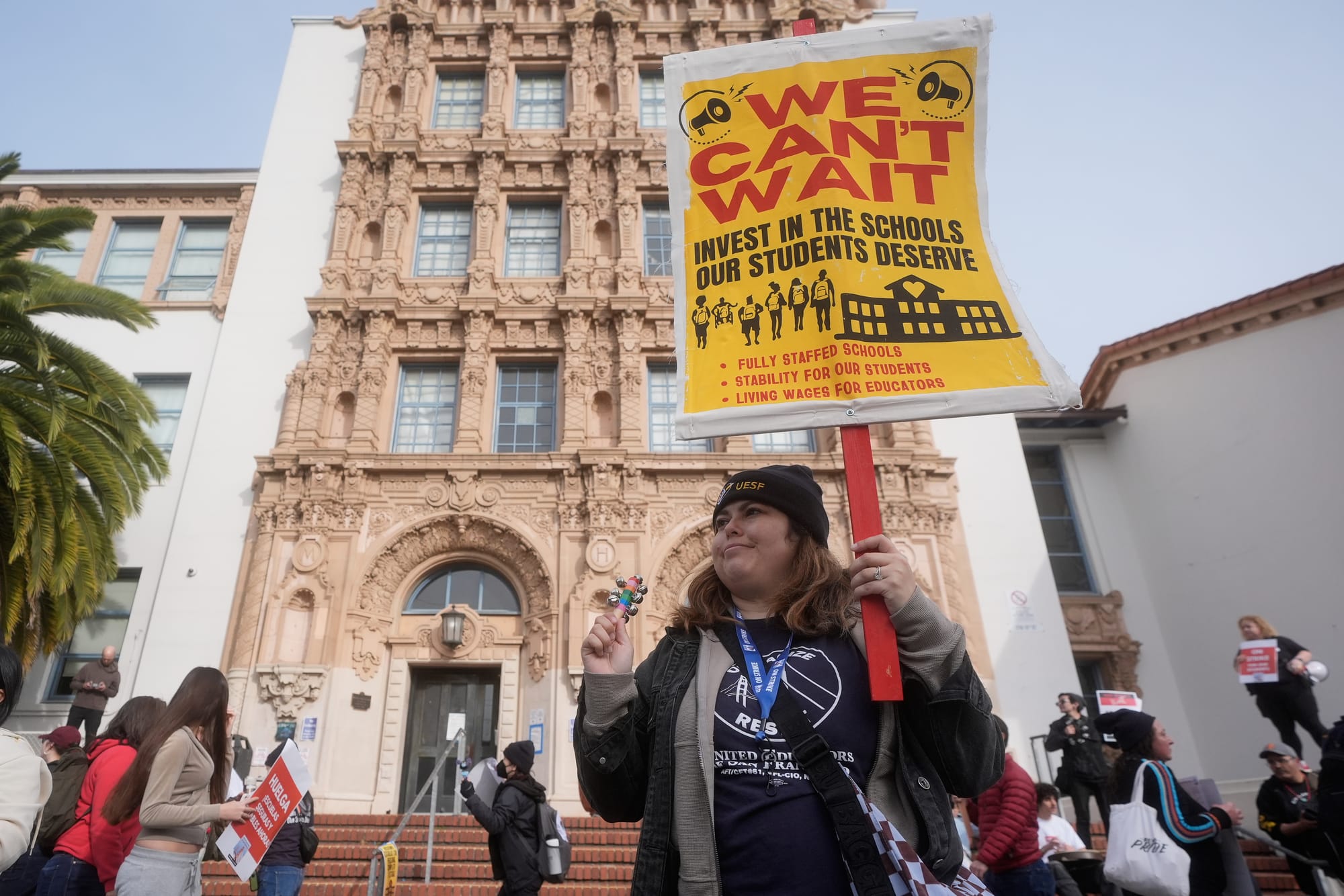 a woman in a jacket and beanie holds a picket sign outside a high school that reads 'we can't wait, invest in the schools our students deserve' with illustrations of students and a school. other teachers and students hold signs around her.