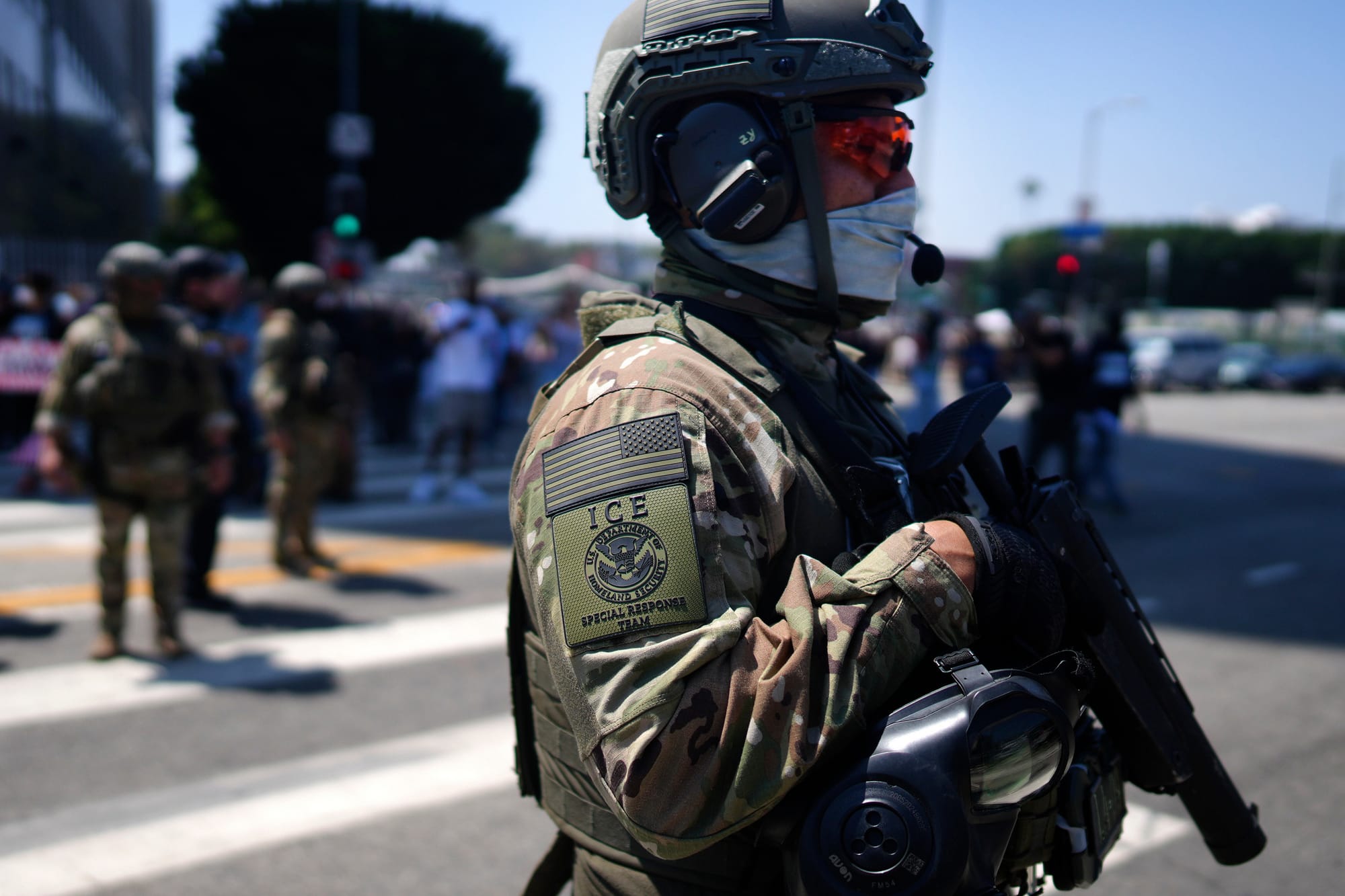 Man in camouflage with a ICE Special Response Team patch on the sleeve (and a backwards USA flag) standing at alert holding a gun