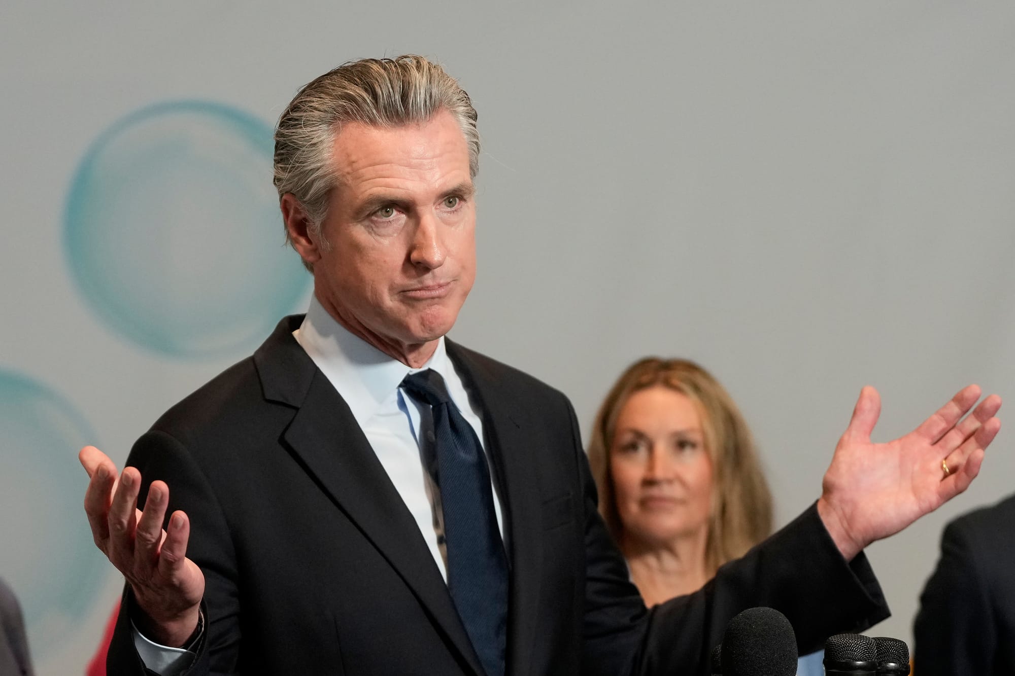 Man with slicked-back grey hair raises his arms in a shrug-like motion at a press conference