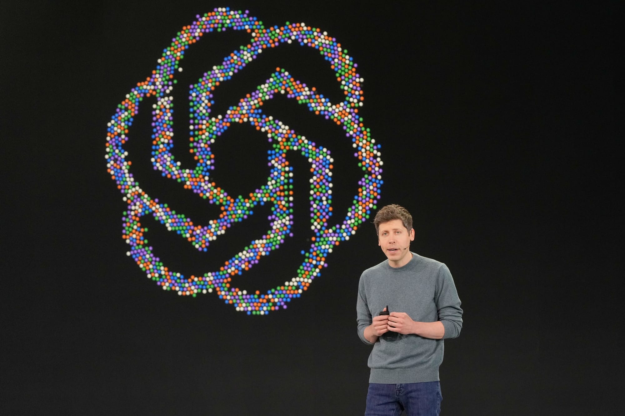 a dorky white man in a gray sweatshirt, Sam Altman, speaks into a headset in front of a logo for Open AI