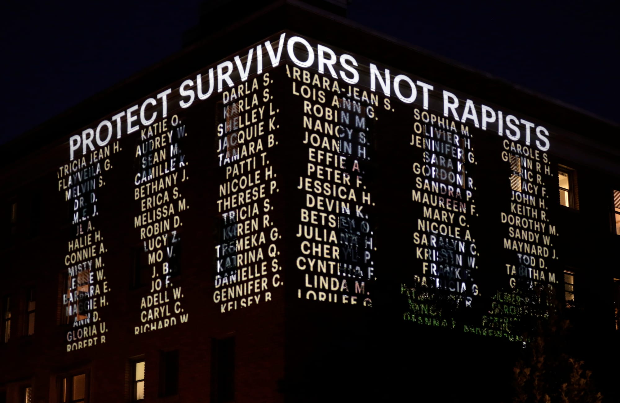 the words 'protect survivors, not rapists' are projected on a building at nighttime, along with around 100 people's first names and last initials in all caps