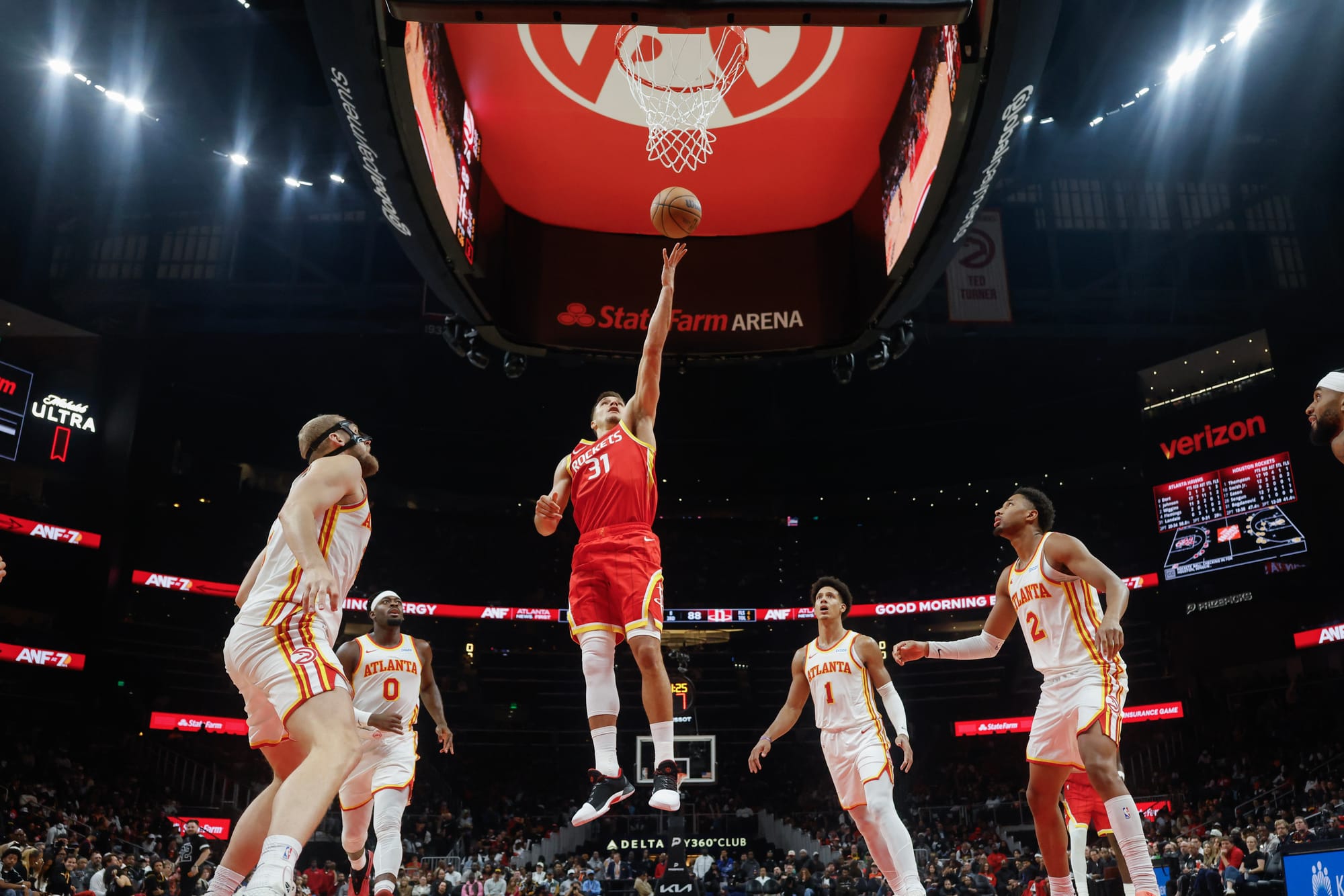 Male basketball player in a red jersey shoots a ball into a hoop while 5 men in white jerseys look on.