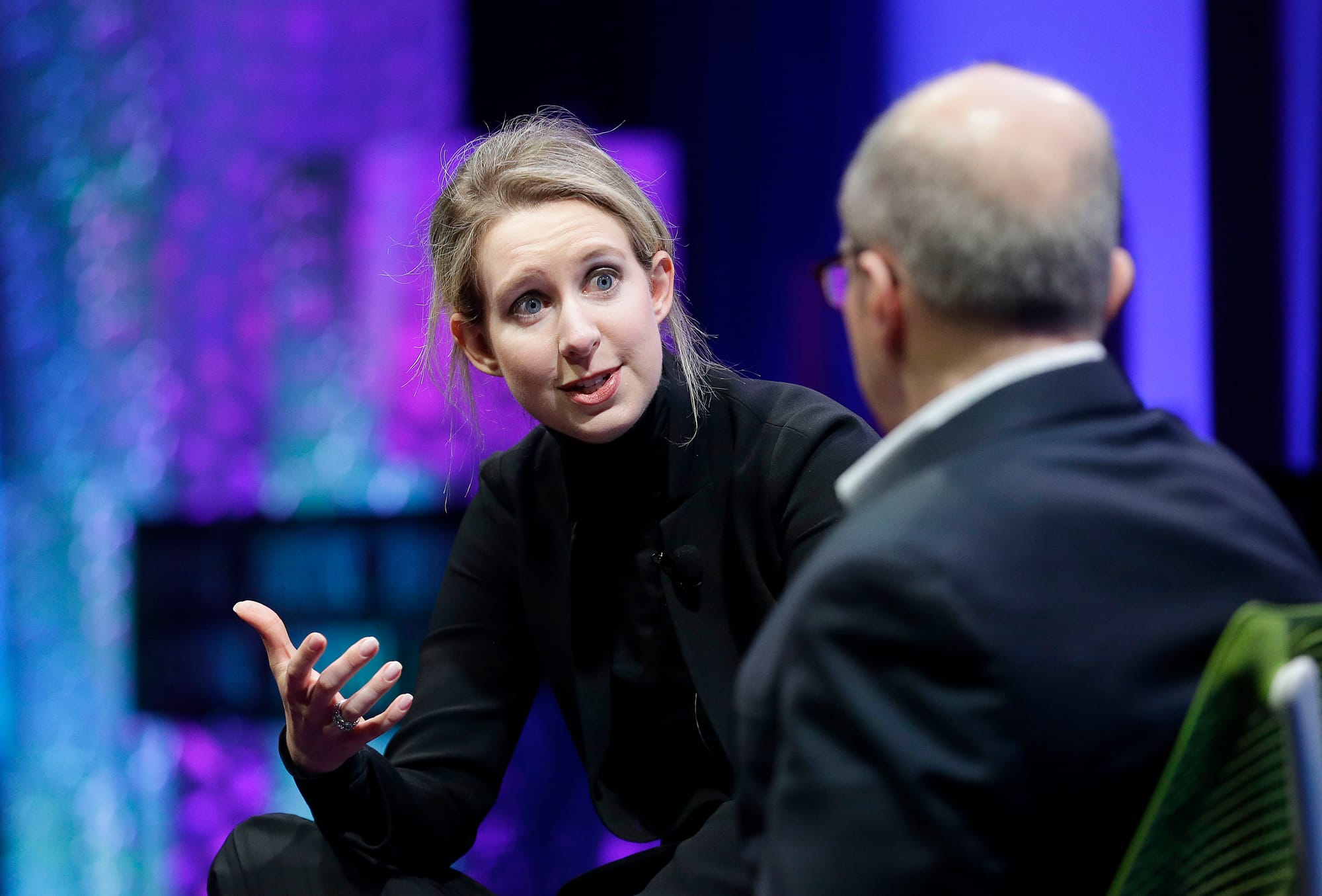 A woman in a black turtleneck gesturing toward a man in a suit.