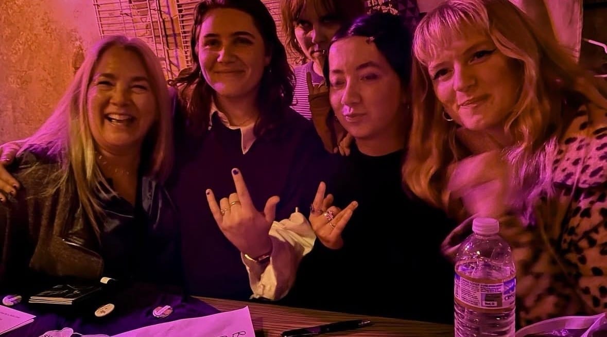 four women pose behind the merch booth at a dimly lit show