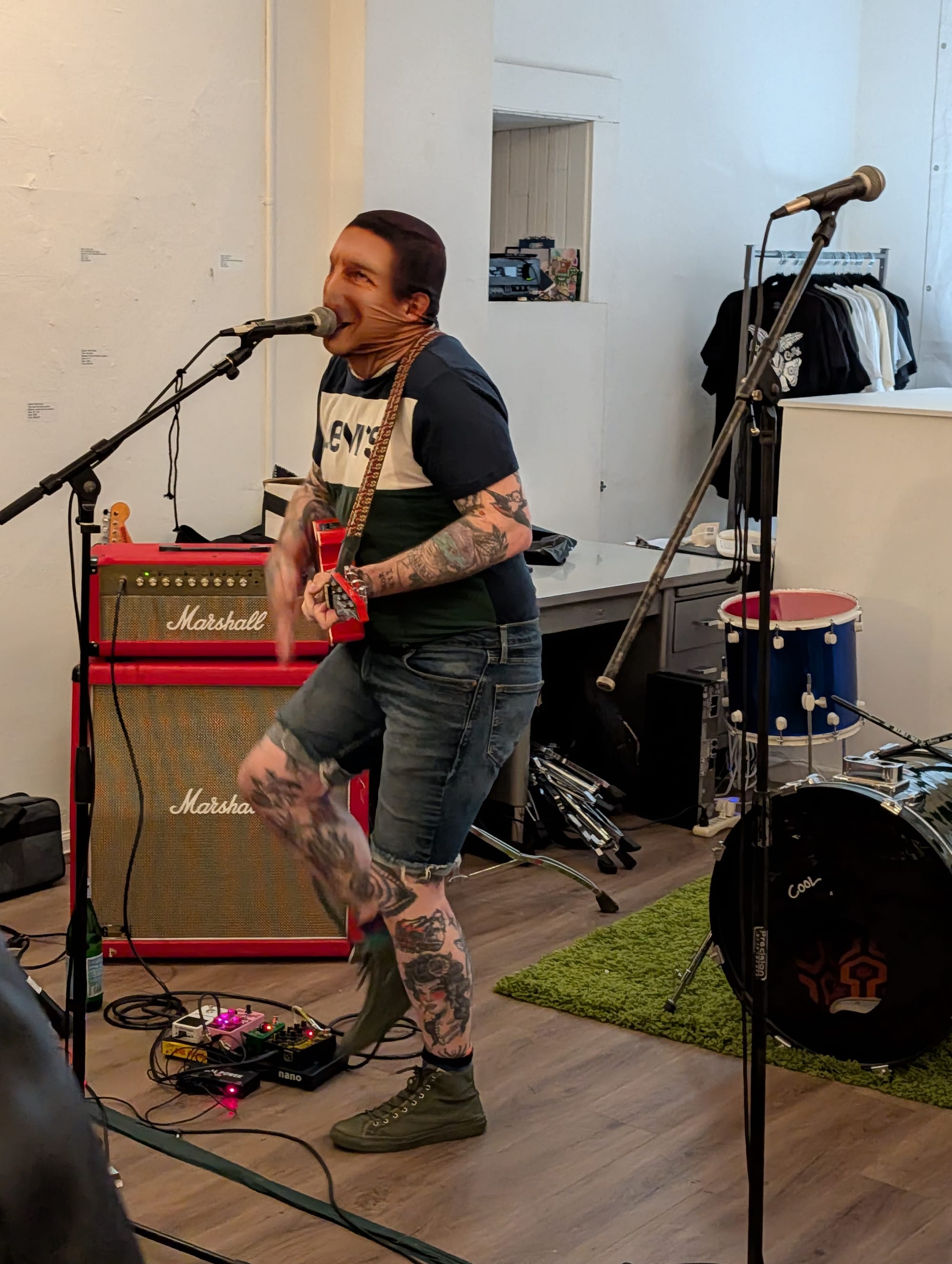 a person with lots of tattoos and a Levi's shirt wearing a screenprinted mask of Daniel Lurie sings and plays the guitar in a gallery show
