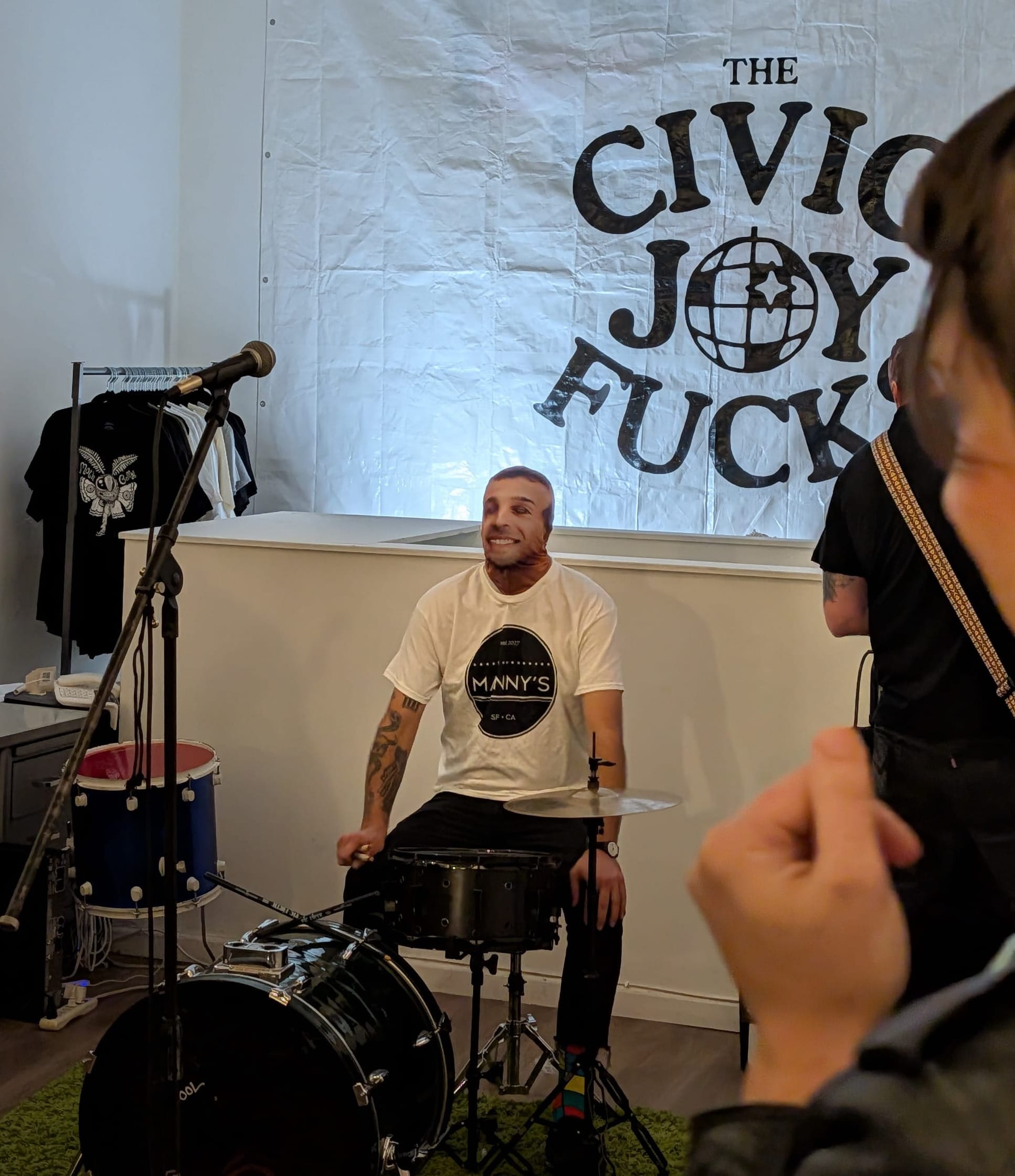 a person in a screen-printed ski mask and a Manny's shirt sits behind a drum kit