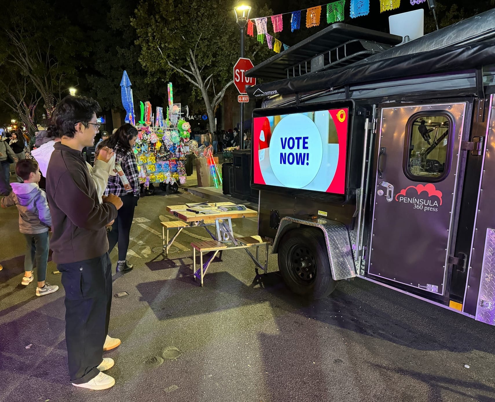 Un hombre está de pie frente a un remolque de color morado que lleva la inscripción «Península 360 Press» en la puerta. Una pantalla grande dice: «¡Vota ahora!».