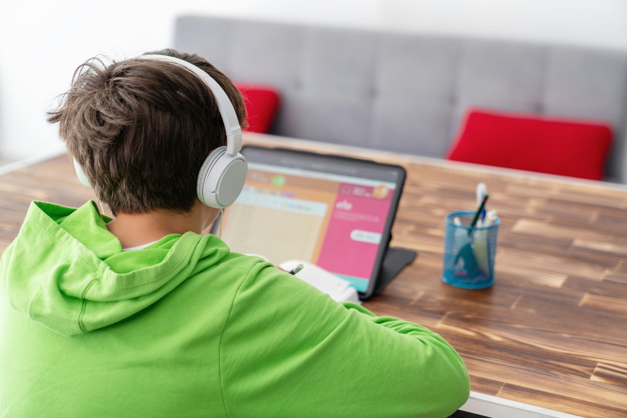 the back of a kid's head as he sits at a table with headphones on looking at a tablet
