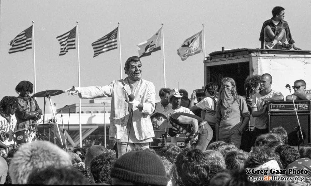 a black and white image shows Jello Biafra with a Ronald Reagan mask on outside the 1984 Democratic National Convention In San Francisco, with a crowd looking on.