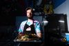 A bearded man in a tie-dyed t-shirt and black apron holds a sheet pan full of meat in an industrial kitchen.