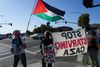 people are seen from behind standing on a street corner holding banners that read 'stop starving gaza' and waving a Palestinian flag