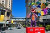 A colorful statue of a bipedal creature with big ears and a huge mouth stands on a red plinth in front of an urban boulevard. A skybridge says "SJSU."