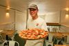 A smiling man in a baseball hat and white t-shirt holds a pepperoni and margherita pizza inside of a restaurant.