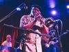 an older Black man plays the trumpet into a microphone on stage, backed by a man on drums and a man playing electric guitar