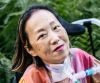 Alice Wong, Asian American woman in a wheelchair with a tracheostomy at her neck connected to a ventilator. She’s wearing a pink plaid shirt and a magenta lip color. 