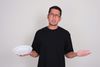 A man in a black shirt and glasses holds an empty white plate in his right hand. He looks directly into the camera with a confused expression.
