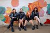 four women in black leather jackets and boots sit on a bench in front of a mural of colorful roses