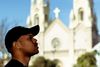 a young Black man in a black ball cap stands in the foreground looking up in front of a church, which is blurry in the background against a blue sky