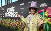 Boots Riley, a Black man in colorful attire and a tall lavender hat, stands on a carpet at a film premiere for 'I Love Boosters,' surrounded by flowers
